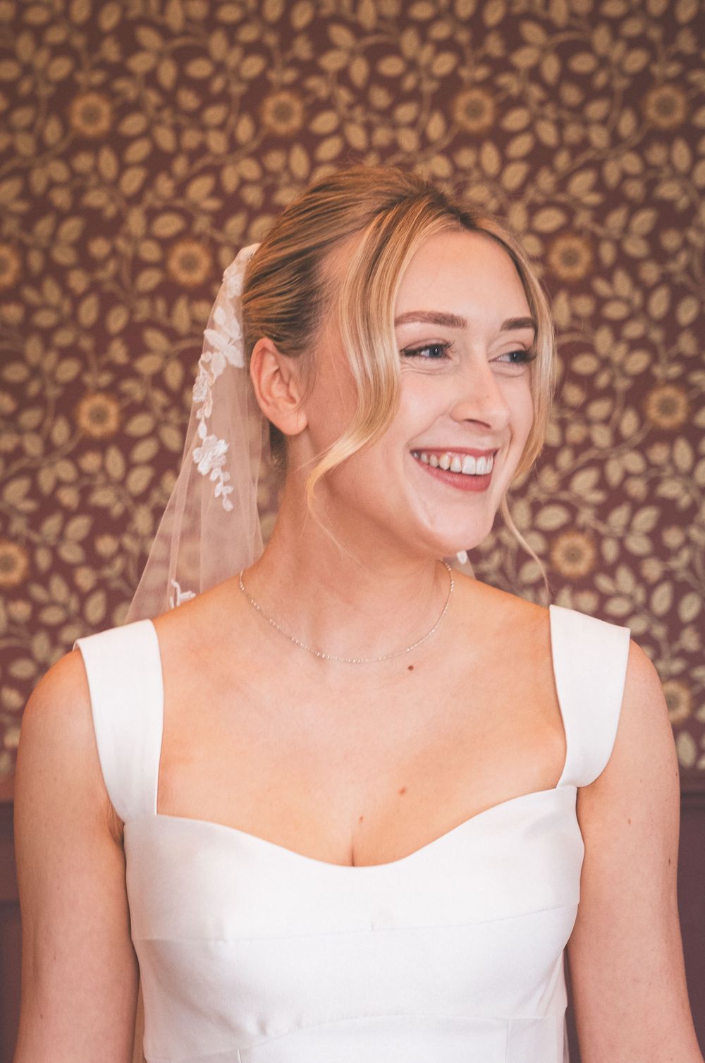 Smiling bride in white dress and floral veil poses against vintage floral wallpaper backdrop.