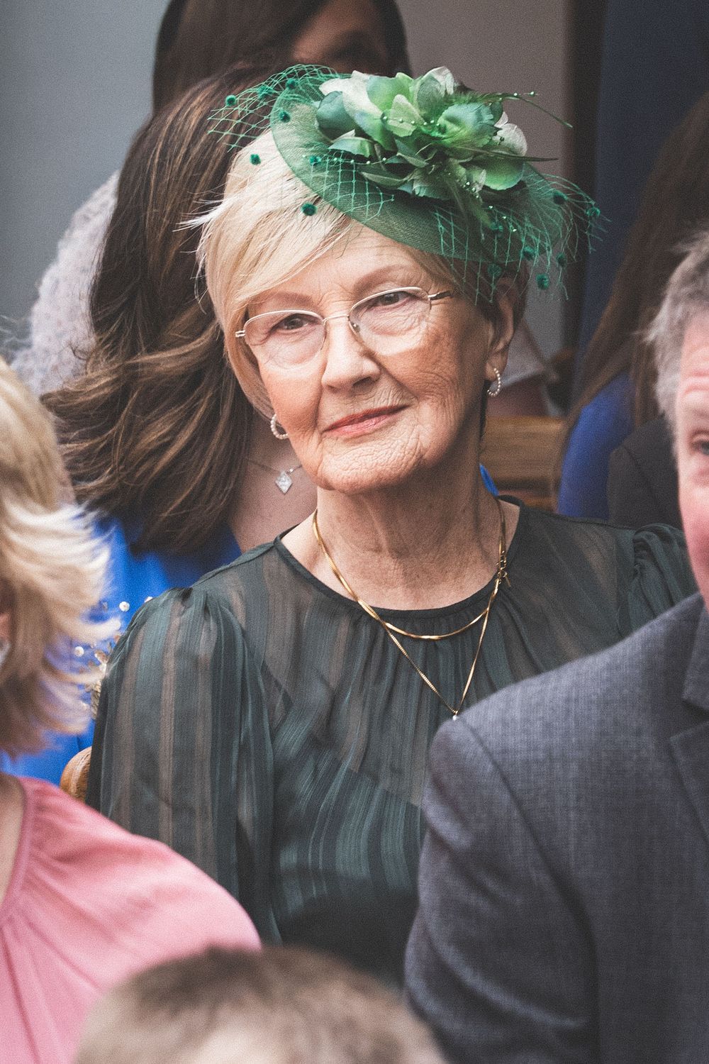 Elegant elderly woman wearing green fascinator hat with floral details and matching dark green blouse at a formal event.