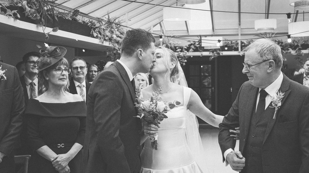 Black and white wedding photo of bride and groom sharing first kiss while guests watch and father of bride looks on.