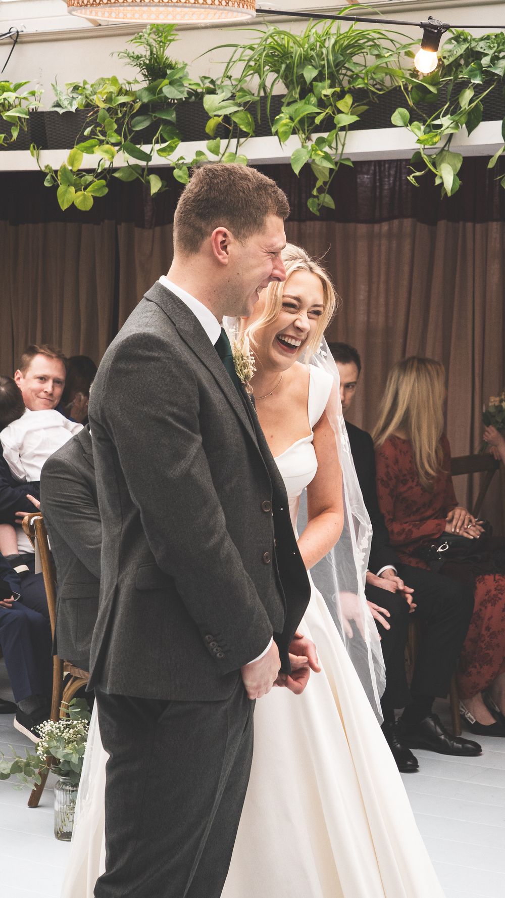 Joyful bride and groom laughing together during their wedding ceremony surrounded by lush greenery decorations.