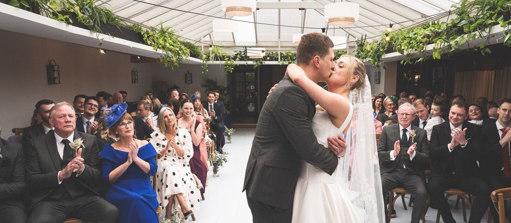 Bride and groom share first kiss at wedding ceremony while guests applaud in elegant venue with greenery.