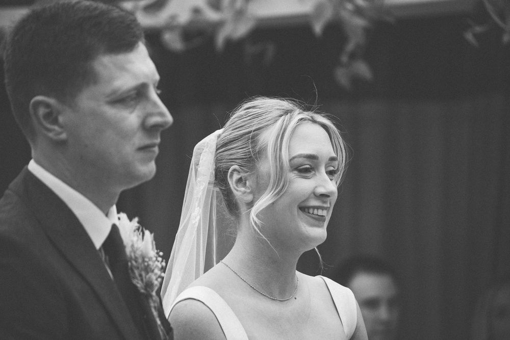 Black and white photo of a smiling bride in veil standing beside groom in suit during wedding ceremony.