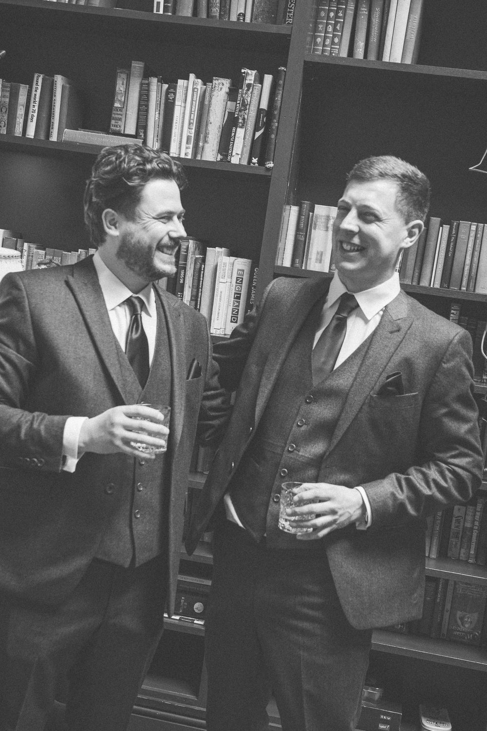 Two men in three-piece suits laugh together holding drinks in front of a bookshelf, black and white photo.