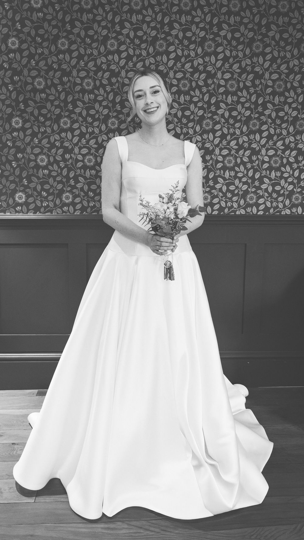 A flowing white wedding gown with a full skirt is showcased against a dark patterned wall.