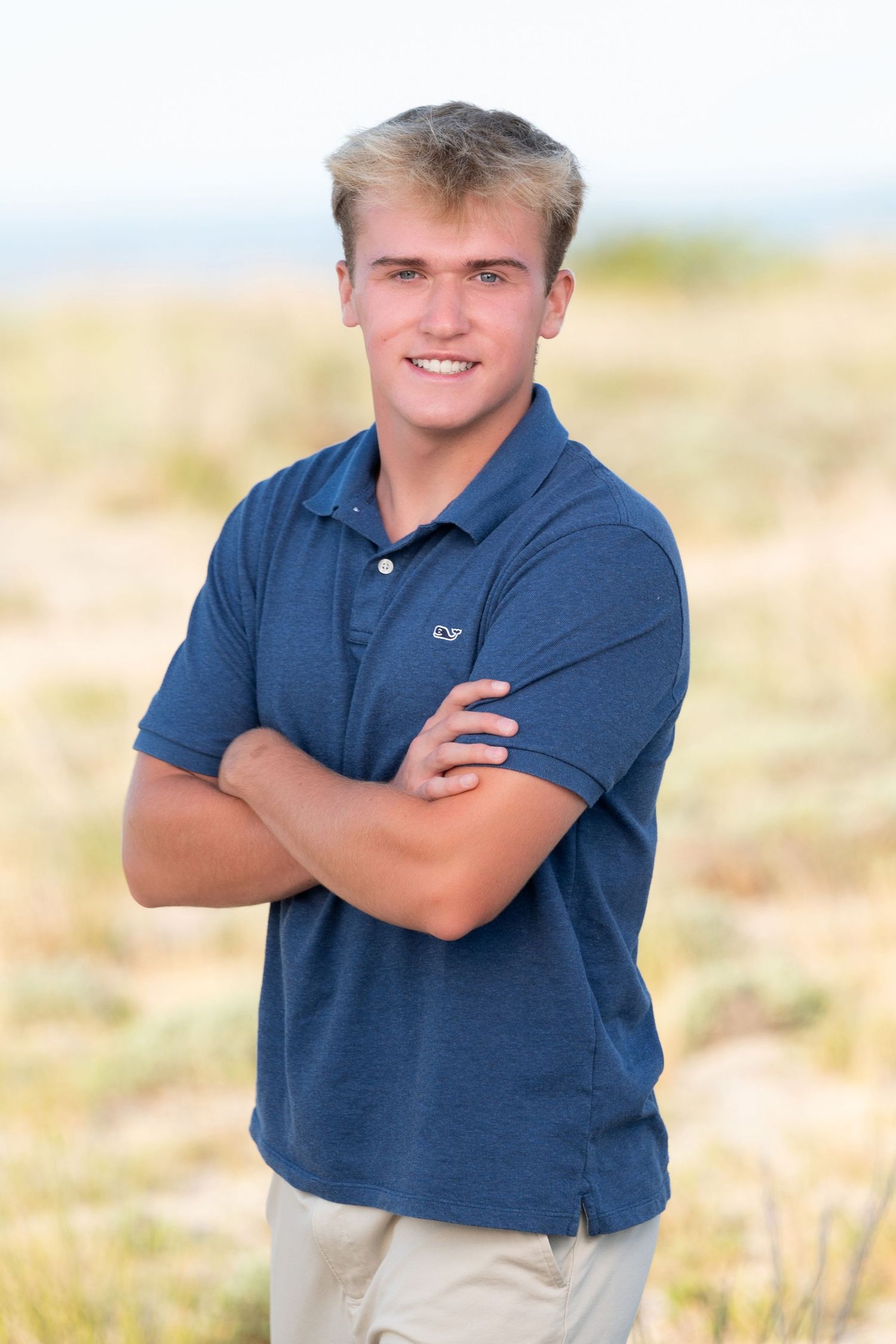 A person in a navy blue polo shirt and khaki pants poses outdoors against a soft blurred natural background.