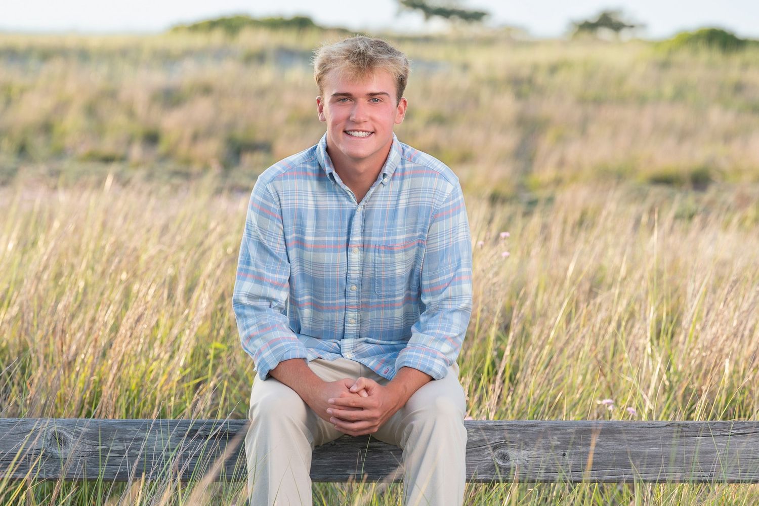 A person in a plaid shirt sits casually on a wooden fence surrounded by tall golden grass at sunset.