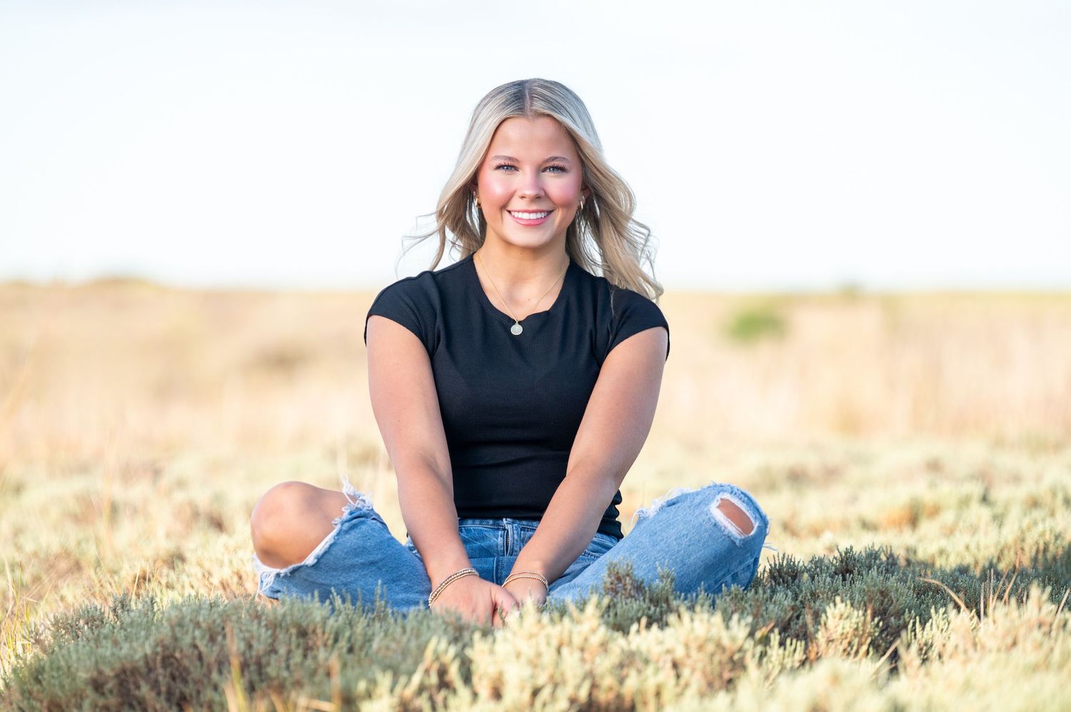 Someone sits cross-legged in a field of golden grass during sunset wearing ripped jeans and a black top.