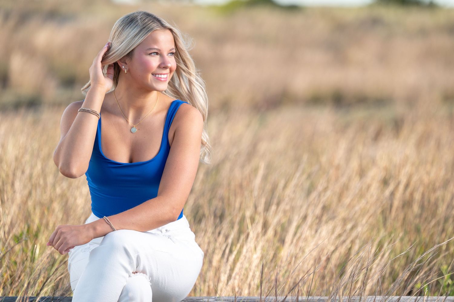 Woman in blue tank top walking through golden wheat field at sunset with sun flare in background.