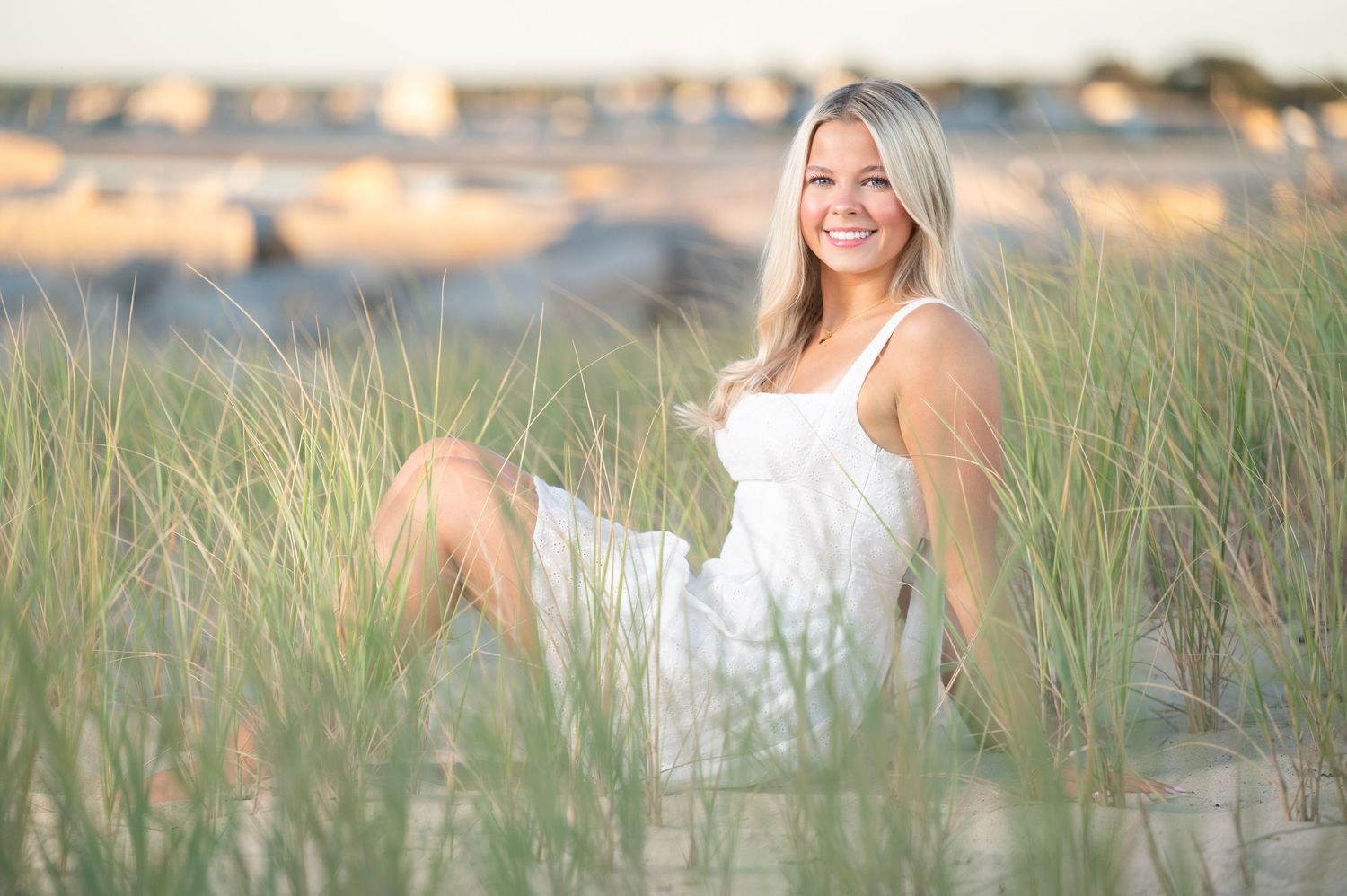 A person in a white dress sits among tall beach grass during golden hour at sunset.