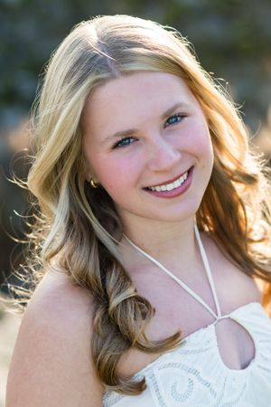 Smiling young blonde woman with blue eyes wearing a white dress and necklace outdoors in natural light.