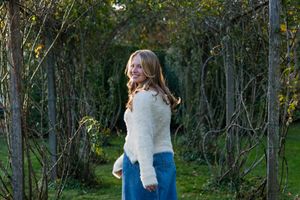 Young woman in white fuzzy sweater and blue skirt smiling outdoors among bare tree branches.