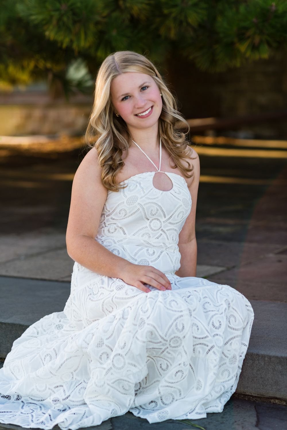 Young blonde woman in white lace formal gown sitting outdoors, smiling brightly for a portrait photo.