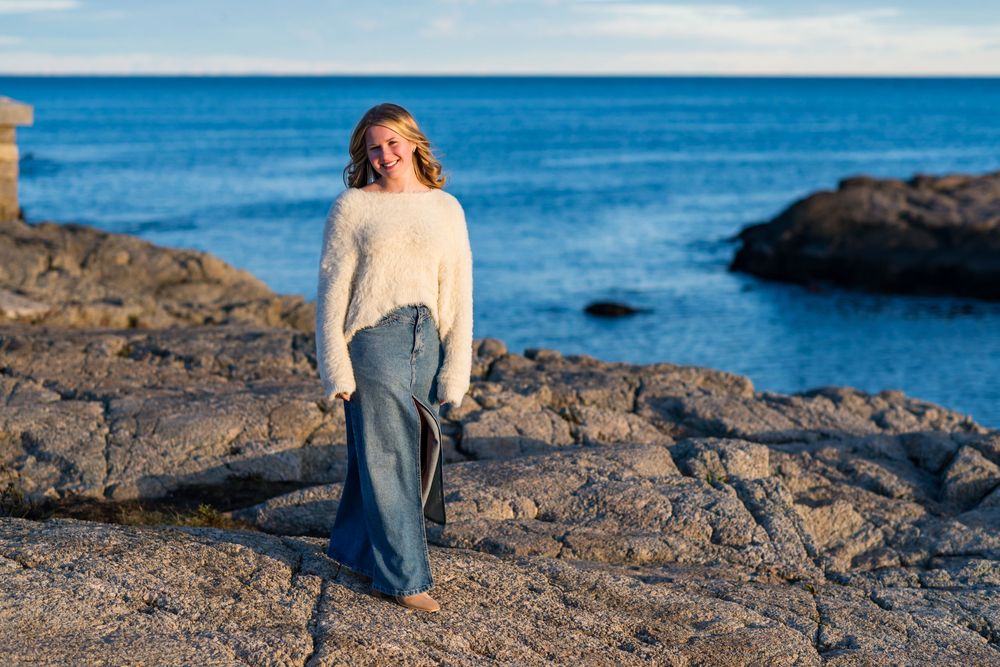Woman in white sweater and denim skirt standing on rocky coastal shore with blue ocean in background.
