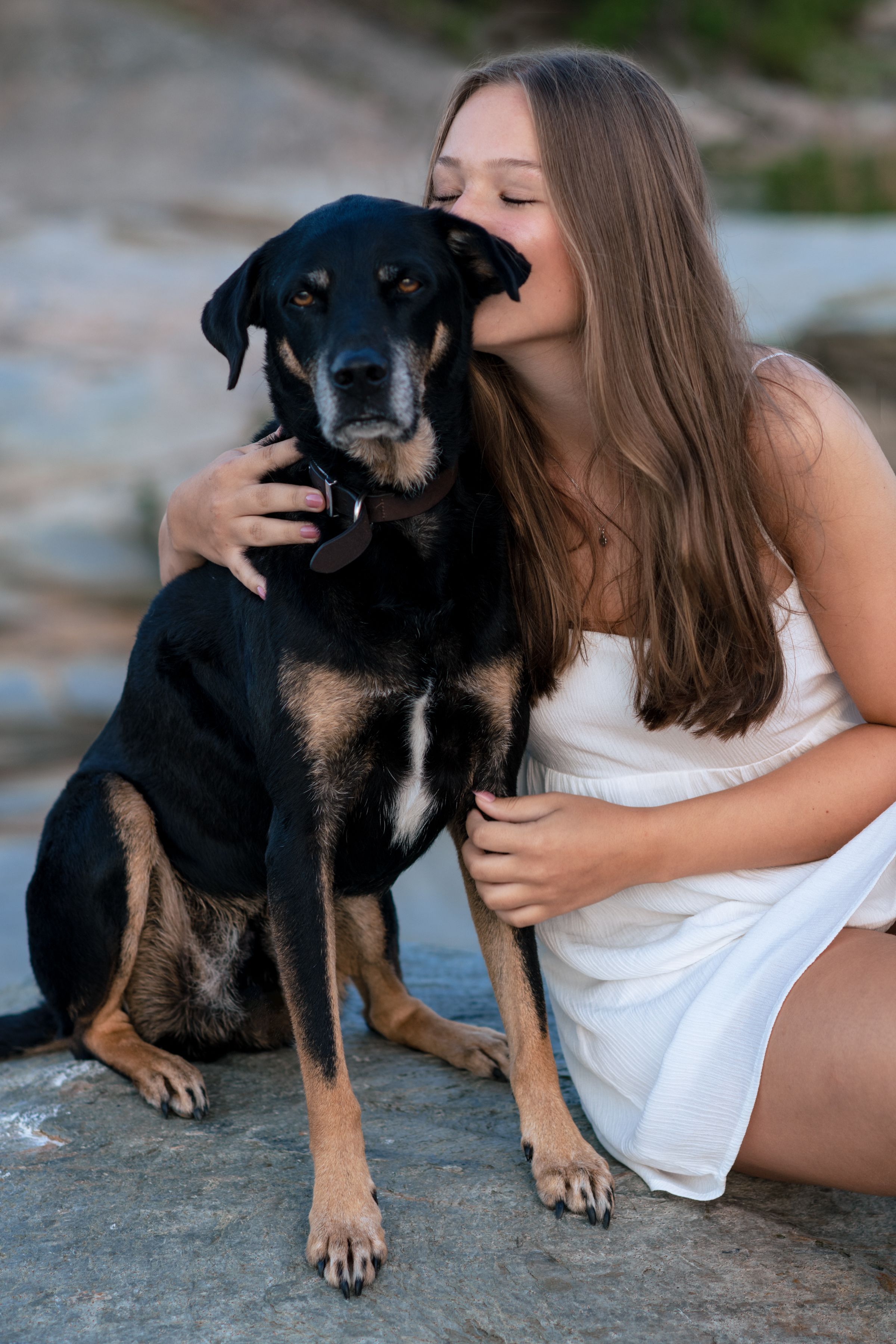 Young woman in white dress kissing her black and tan dog while sitting on rocks outdoors.