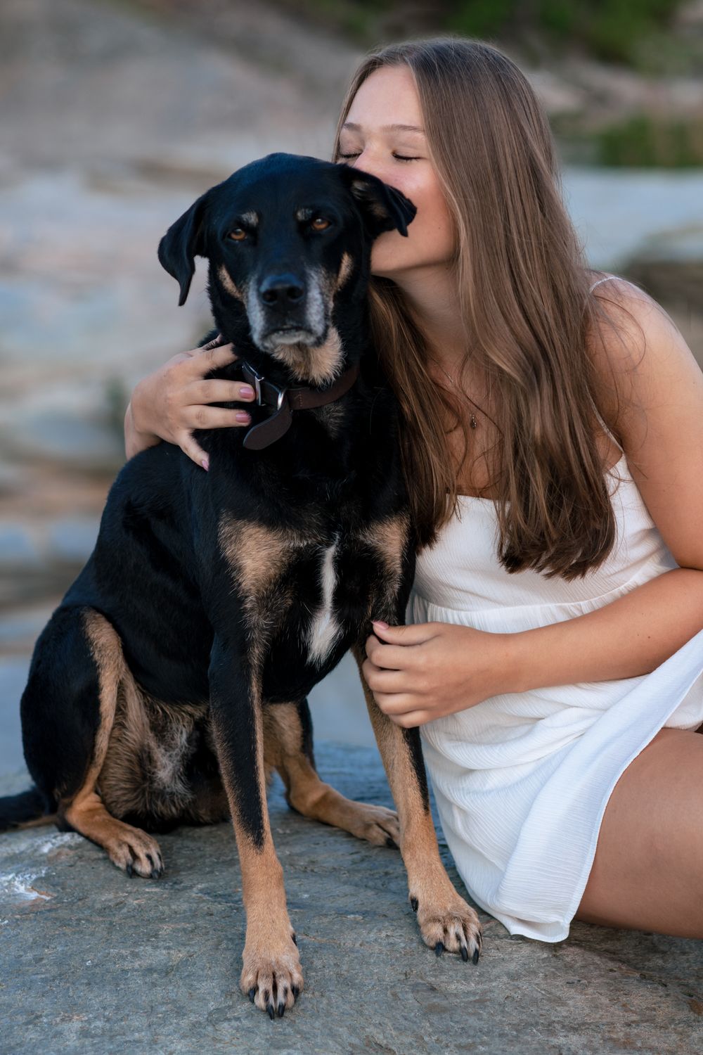 Young woman in white dress kissing her black and tan dog while sitting on rocks outdoors.