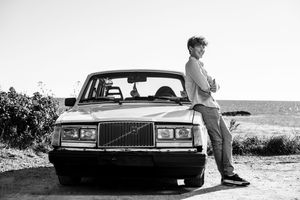 Young man leaning against a classic Volvo car near the ocean in a black and white photo.