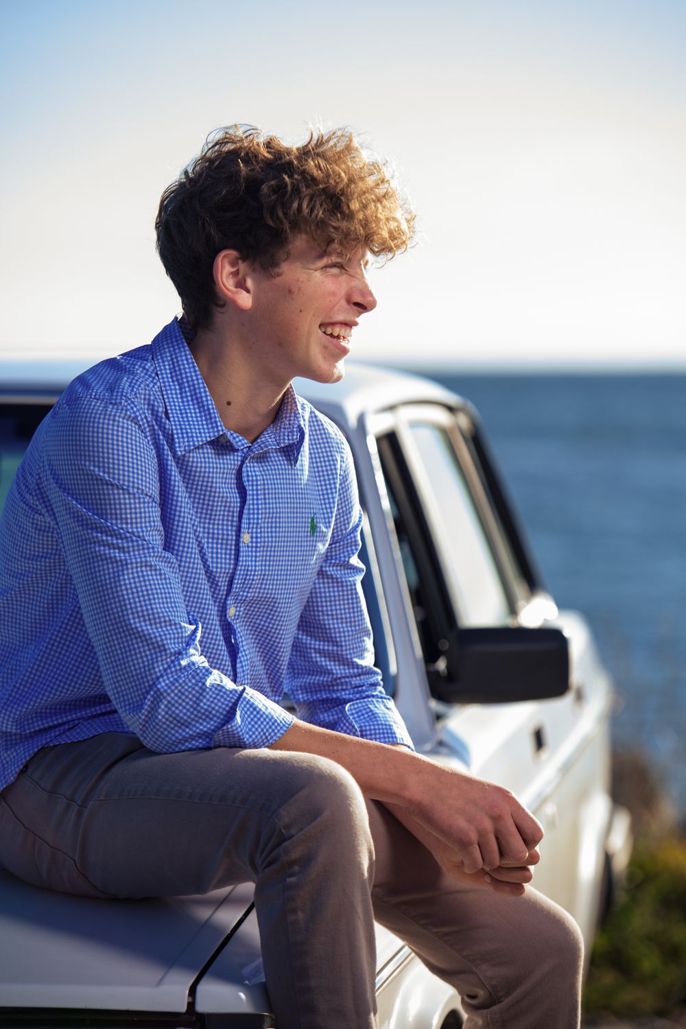 Young man with curly hair in blue shirt smiling while sitting on white car hood near ocean.