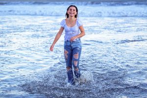Young woman in ripped jeans and white top laughing while splashing through ocean waves on a sunny beach.