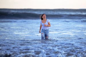 A young woman in ripped jeans and white top laughs joyfully while standing in ocean waves at the beach.