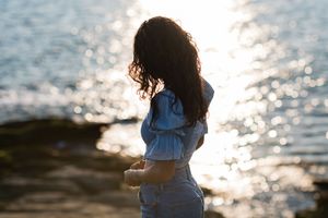 Young woman with curly hair standing by the shimmering sea at sunset, wearing a blue top and jeans.