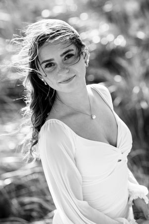 Black and white portrait of a smiling bride in a white dress with pearl necklace, hair blowing in the wind outdoors.