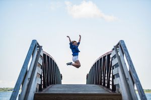 A joyful child jumps with arms raised on a wooden bridge overlooking a calm lake under a bright blue sky.