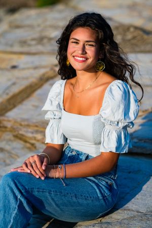 Young woman with dark curly hair smiles outdoors, wearing white crop top, jeans, and gold earrings, seated on stone.