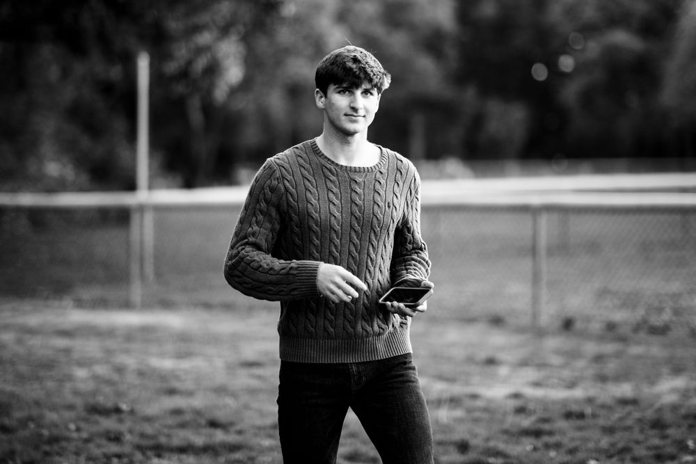 Young man in cable-knit sweater and dark jeans standing outdoors in black and white portrait photo.