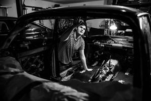 Black and white photo of a woman working on a vintage car in a garage, illuminated by a single overhead light.