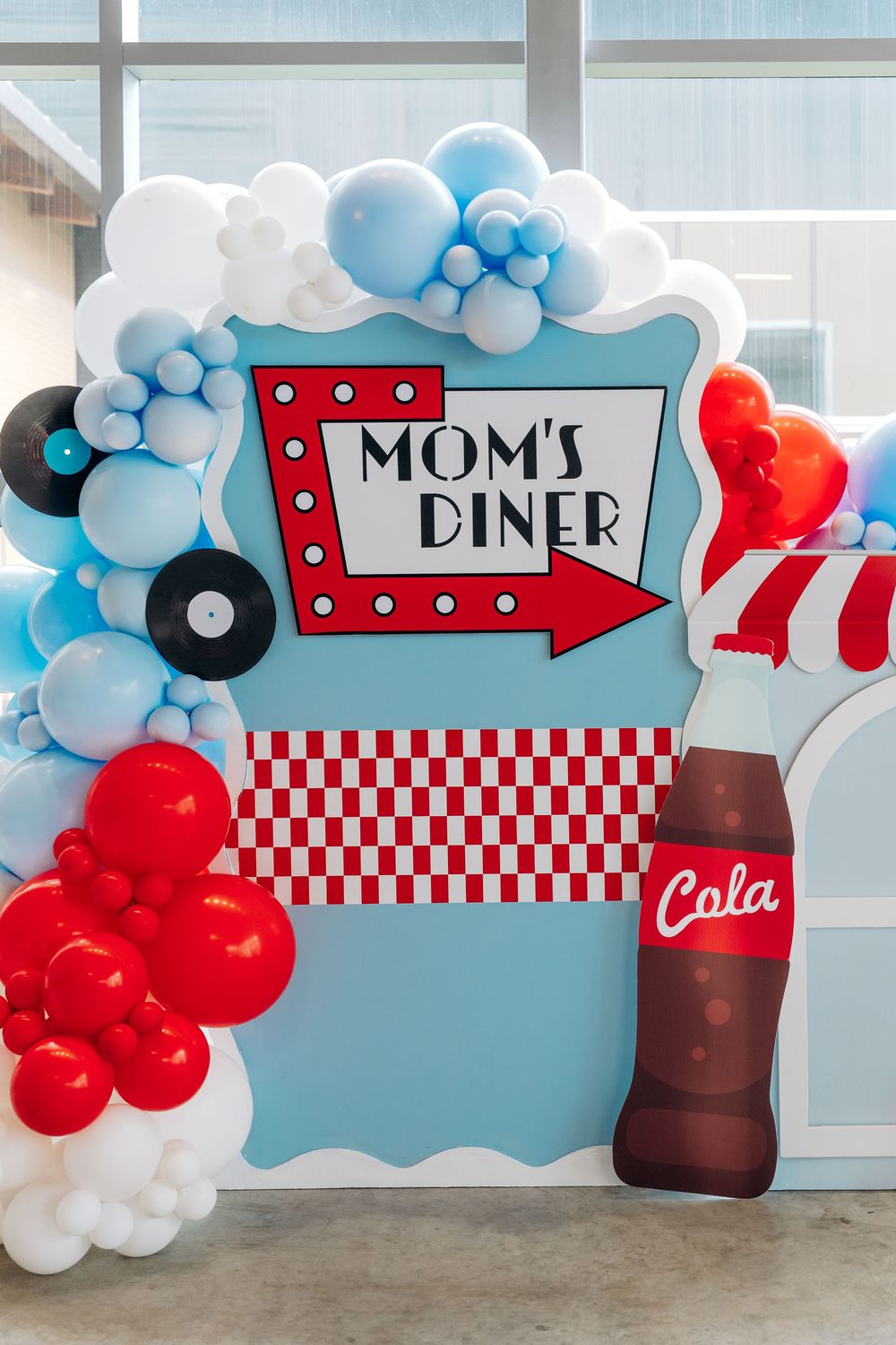 Retro diner themed party backdrop with balloon arch and Mom's Diner sign in red and light blue colors.