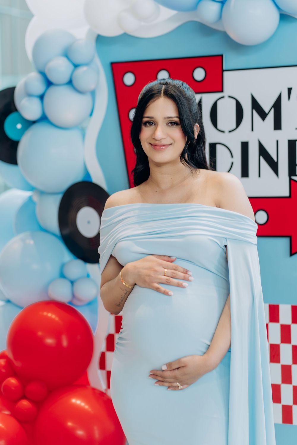 A maternity photoshoot against a backdrop with blue balloon clouds and a retro diner sign with checkered pattern.