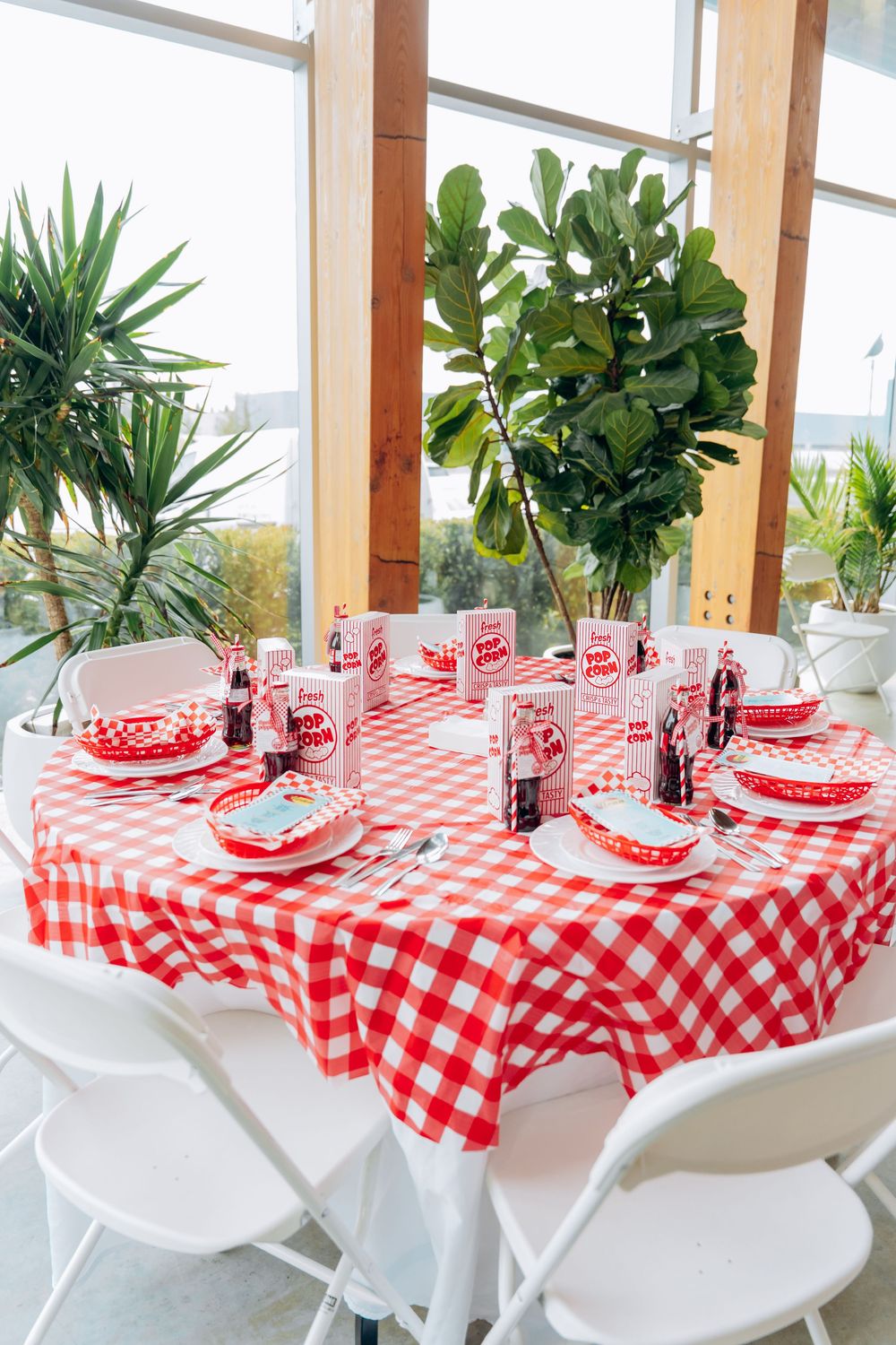 Table set with red and white checkered tablecloth, vintage-style place settings, and indoor plants by sunlit windows.