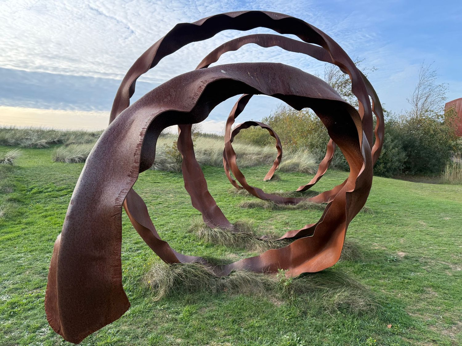 Large rusty Corten steel spiral sculpture displayed in an open green meadow under a blue sky.