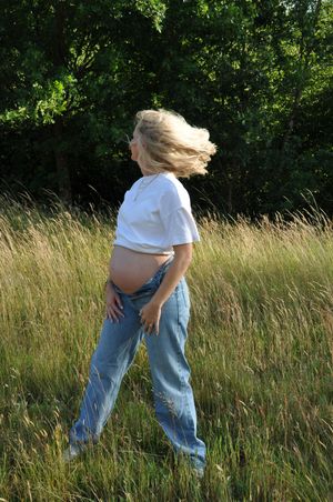 Pregnant blonde woman in white t-shirt and jeans walking through a sunny meadow with hair blowing in the wind.