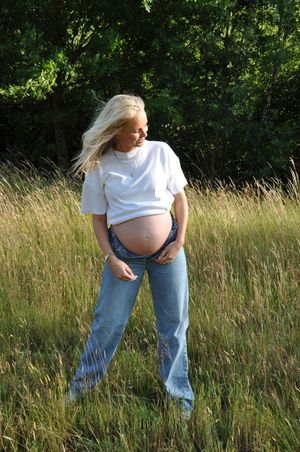 Pregnant blonde woman in white t-shirt and jeans standing in tall grass field, looking down at her baby bump.