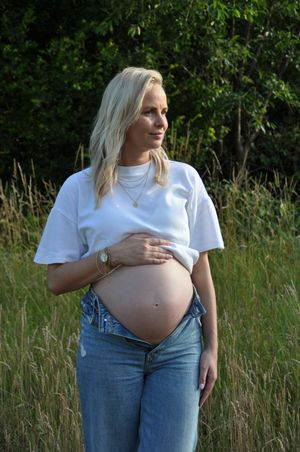 Pregnant blonde woman holding baby bump in white tee and jeans in lush meadow.