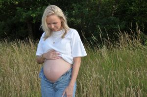 Pregnant blonde woman in white tee and jeans cradling baby bump in golden meadow.