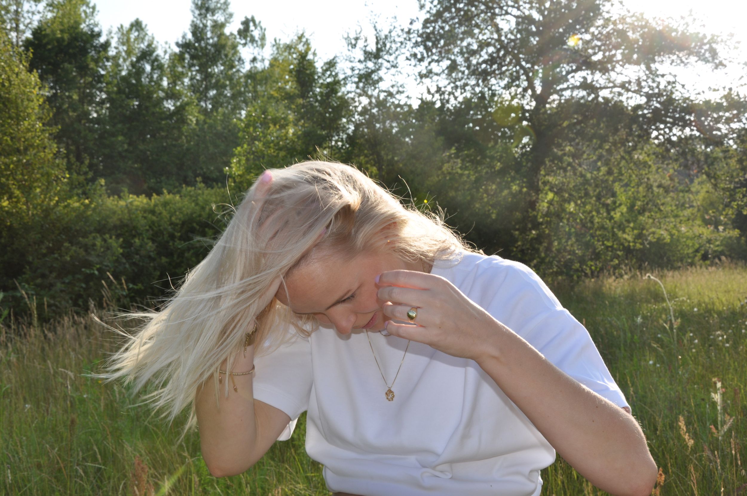 Blonde woman tossing hair in sunlit green field wearing white t-shirt.