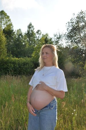 Pregnant blonde woman in white tee holding bump while gazing in sunny meadow.