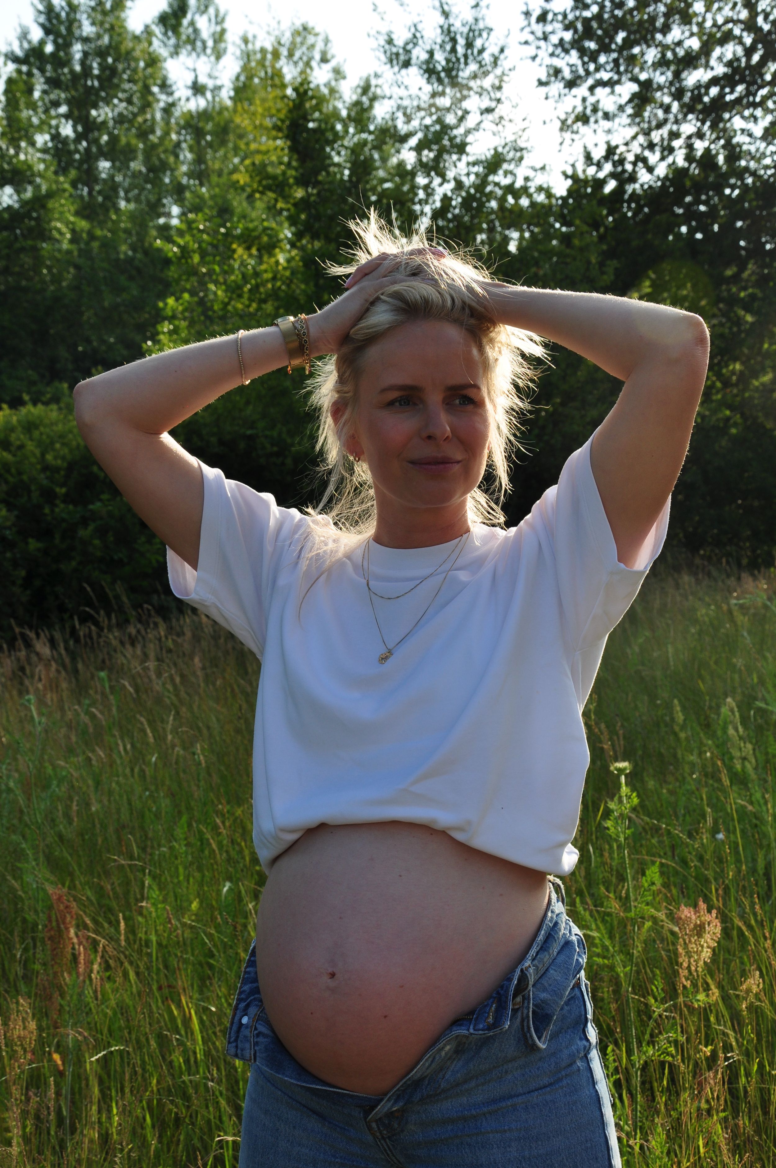 Pregnant blonde woman in white t-shirt and jeans posing outdoors in a field, lifting shirt to reveal baby bump.