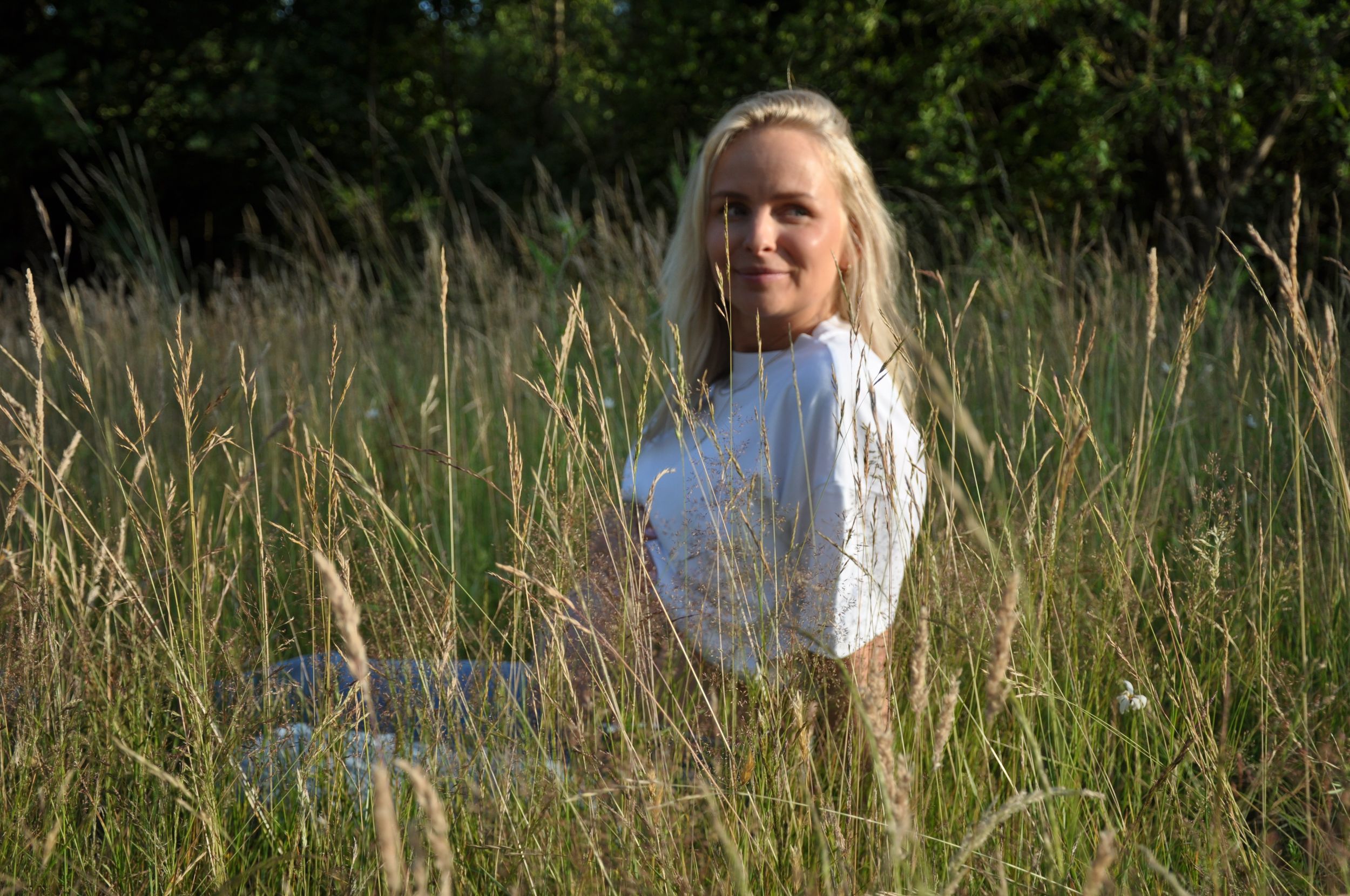 Smiling blonde woman sitting in tall green grass field on a sunny day.