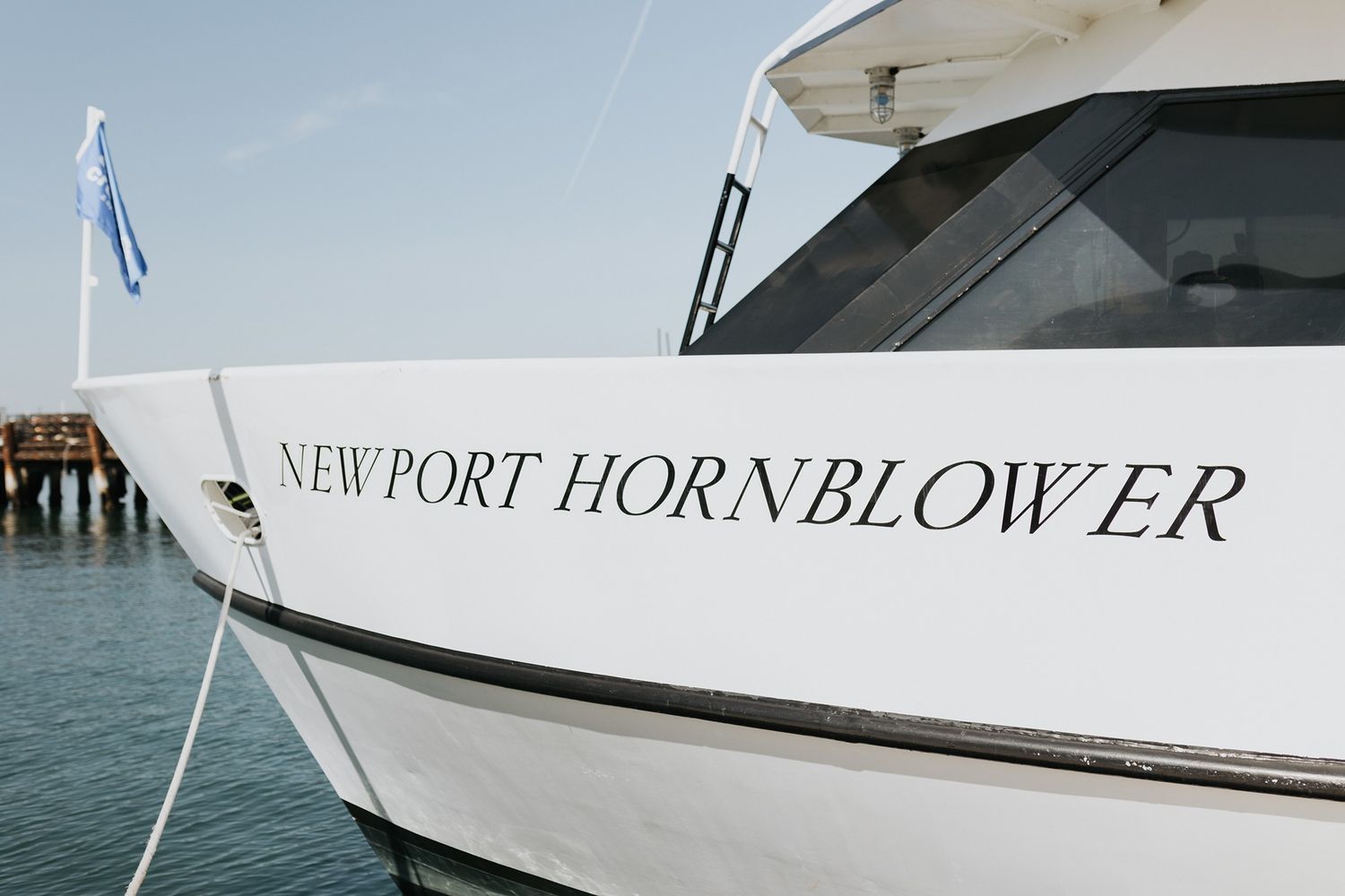 White boat named Newport Hornblower docked at a marina with calm waters and clear skies in the background in San Diego.