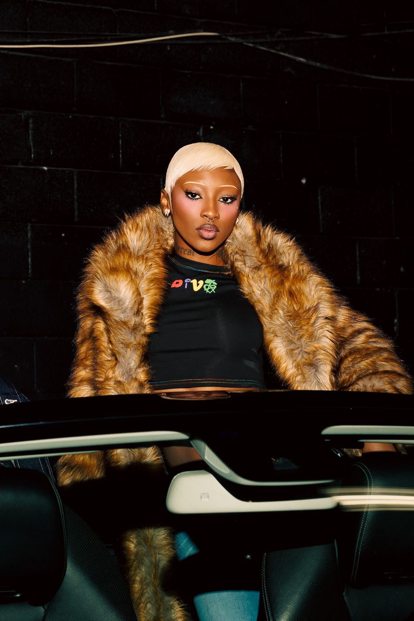 A person wearing a black top and fur-trimmed coat sits in a darkly lit car interior.