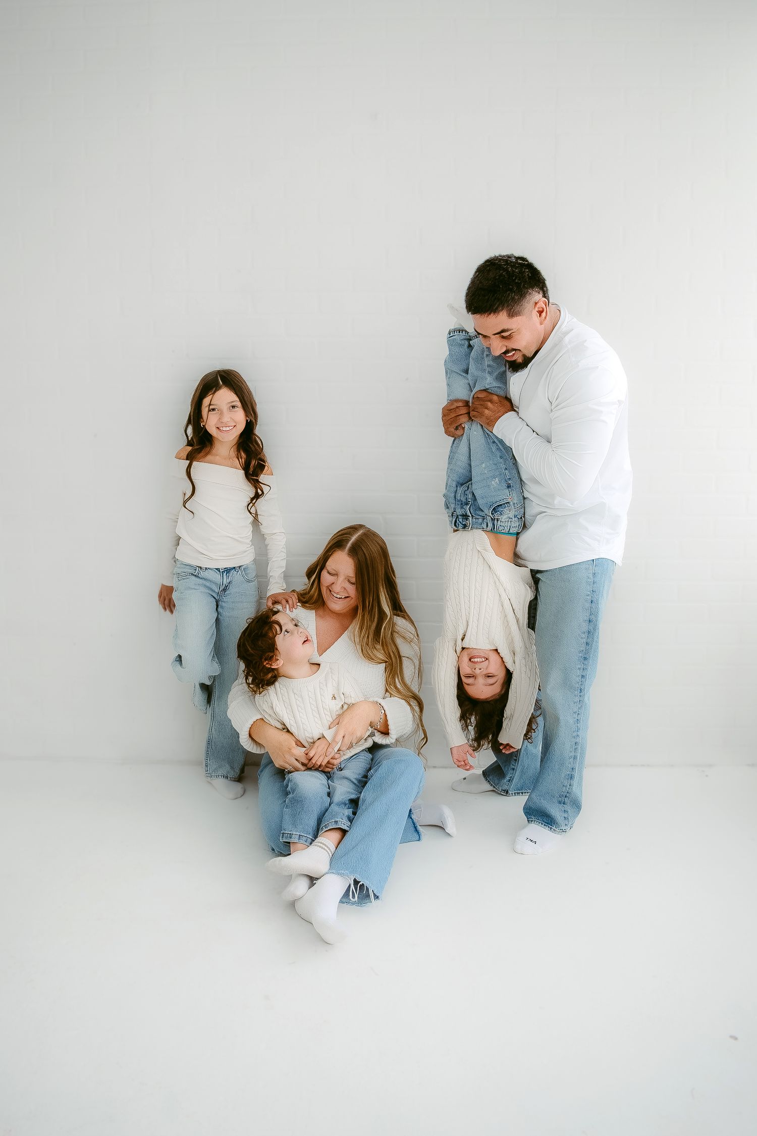 Family wearing white and denim lounging playfully together against white backdrop in casual lifestyle photoshoot.