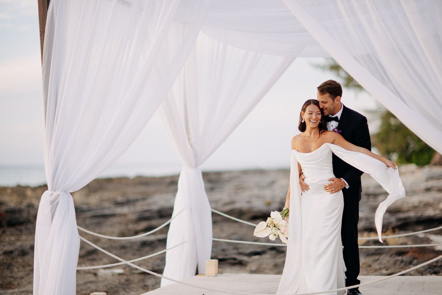 Flowing white fabric drapes create an ethereal beach wedding backdrop against a rocky coastal shoreline.