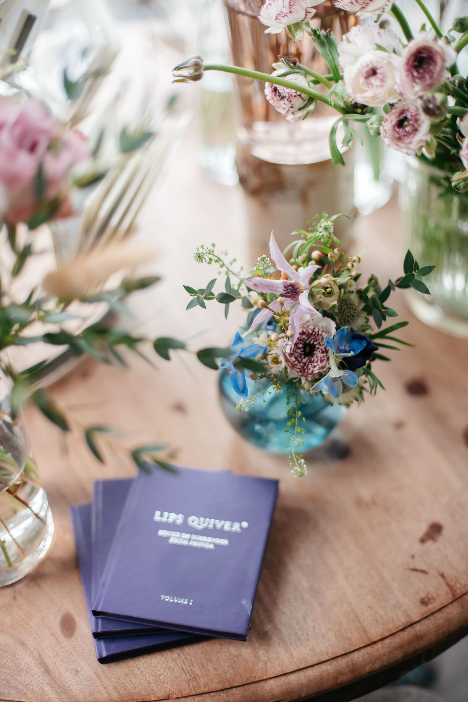 Small blue glass vase with delicate pink flowers and greenery sits beside purple books on rustic wooden table.