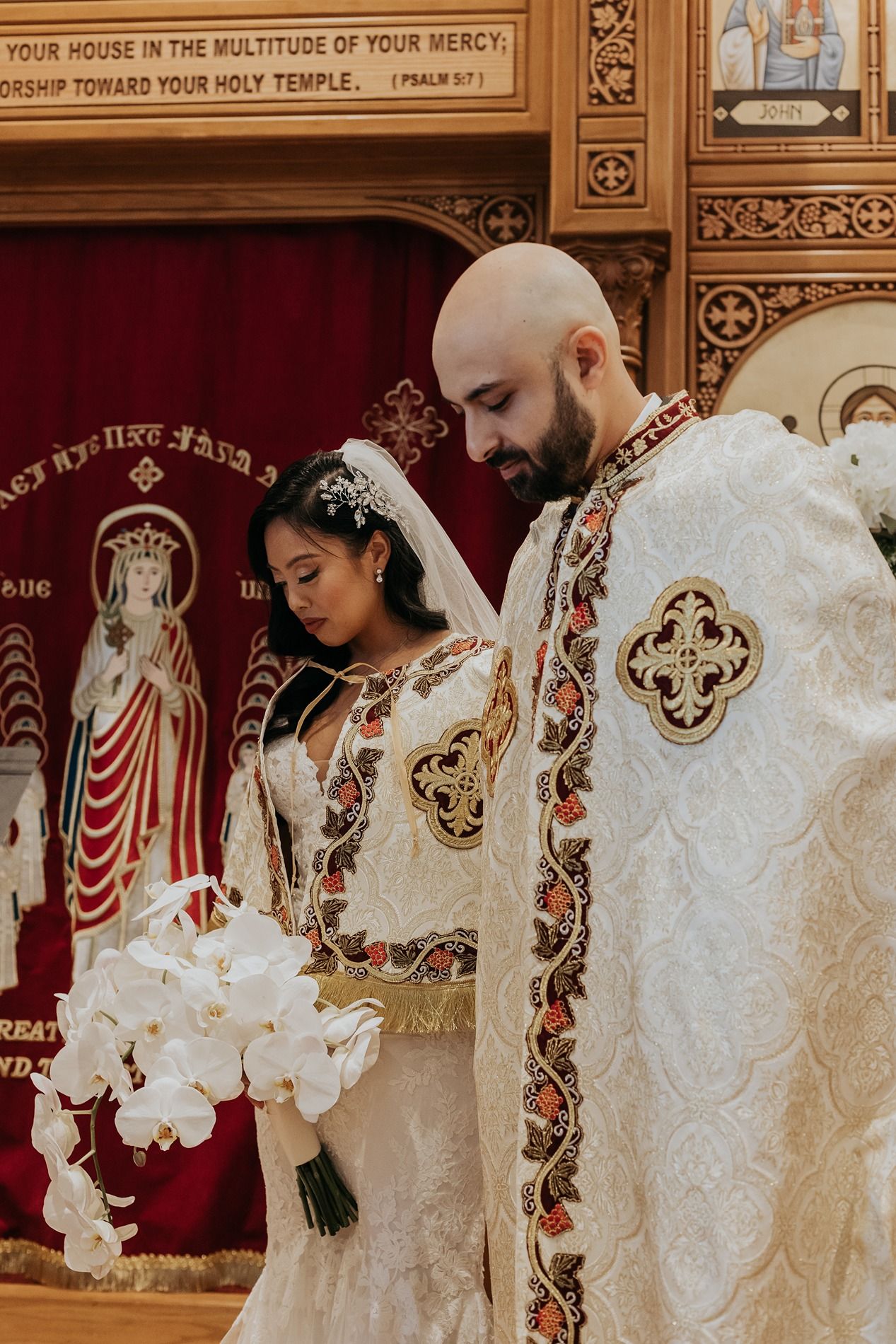 Couple in white traditional wedding attire standing together during an ornate church ceremony with decorative walls.