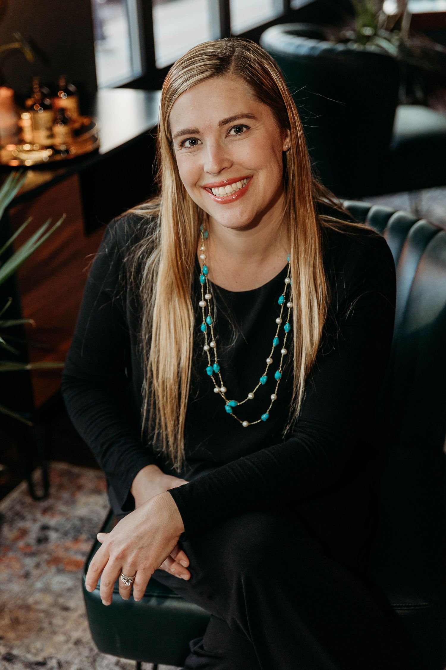 Professional portrait of someone wearing a black top and turquoise beaded necklace while sitting on a dark leather chair.