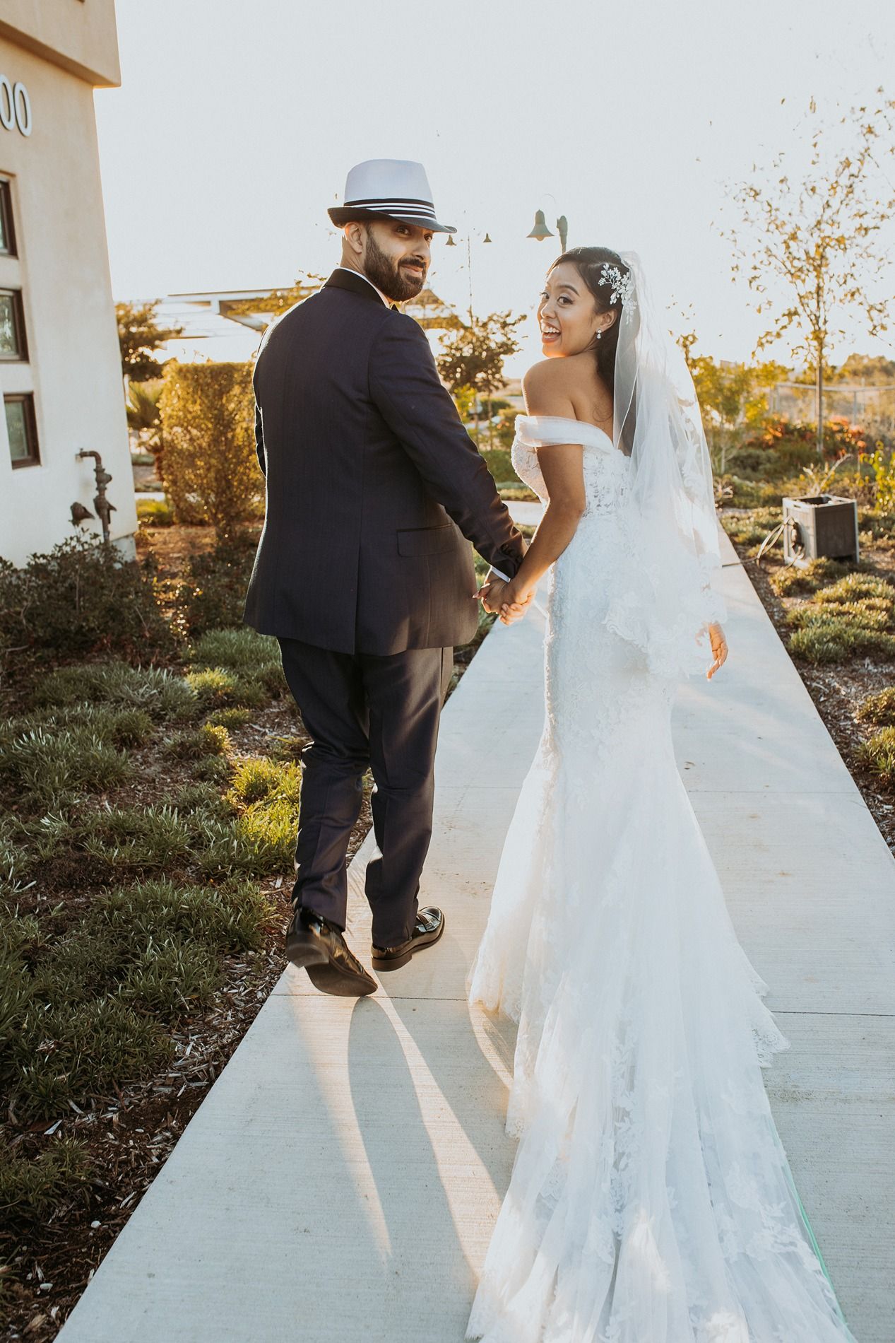 A bride in a white off-shoulder gown and groom in dark suit share a joyful moment during their outdoor sunset wedding.