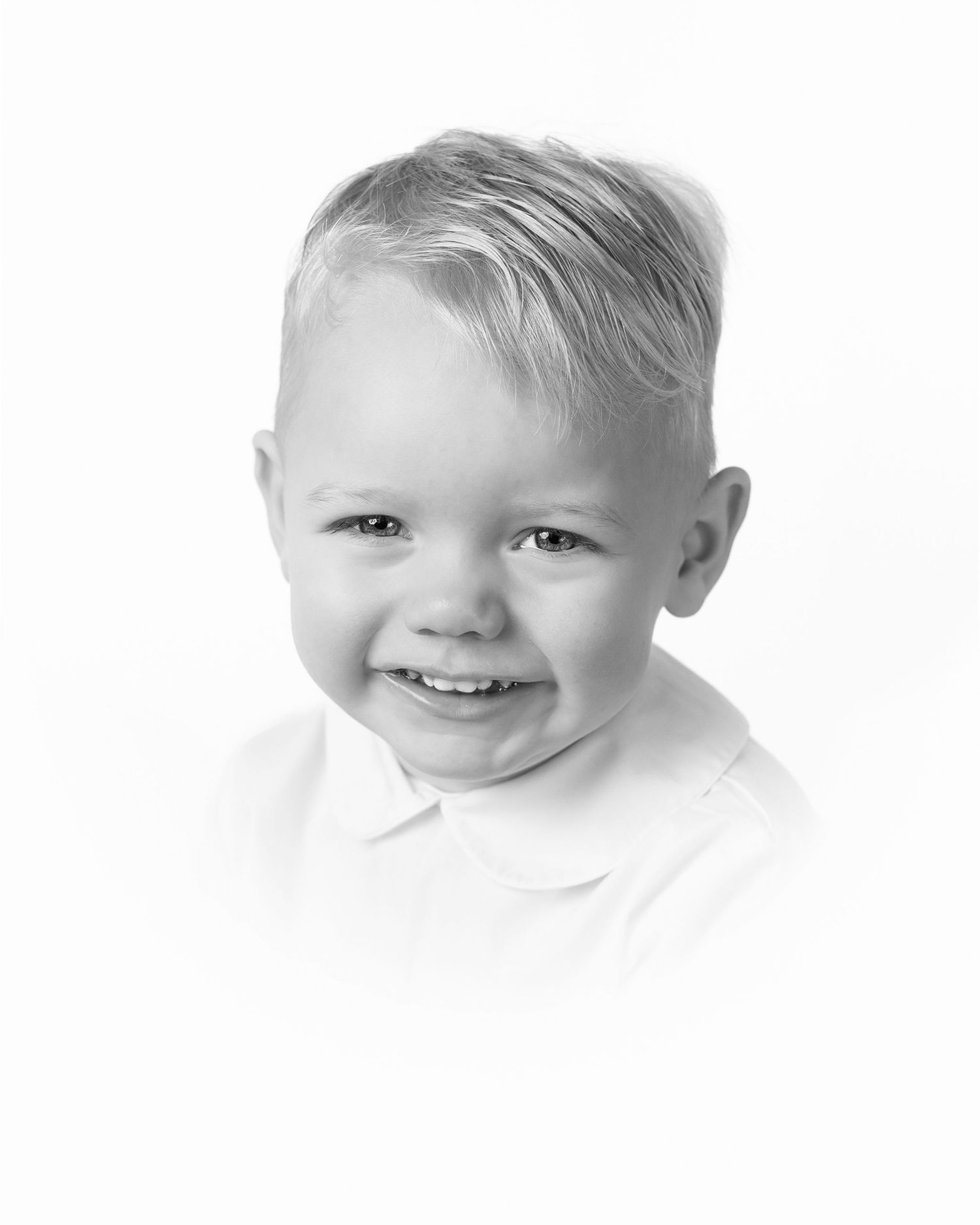A young child with blonde hair wearing a white shirt smiles brightly against a white background.